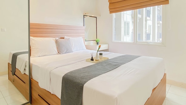 A bedroom with a bed, bedside table, mirror, window with blinds, and a view of buildings outside. - Practical 2Br For 4 Pax At Podomoro Golf View Apartment (Gunung Putri)