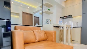 A modern living room with a sofa, a dining area with a chair, a kitchen with cabinets, and a built-in oven. - Practical 2Br For 4 Pax At Podomoro Golf View Apartment (Gunung Putri)