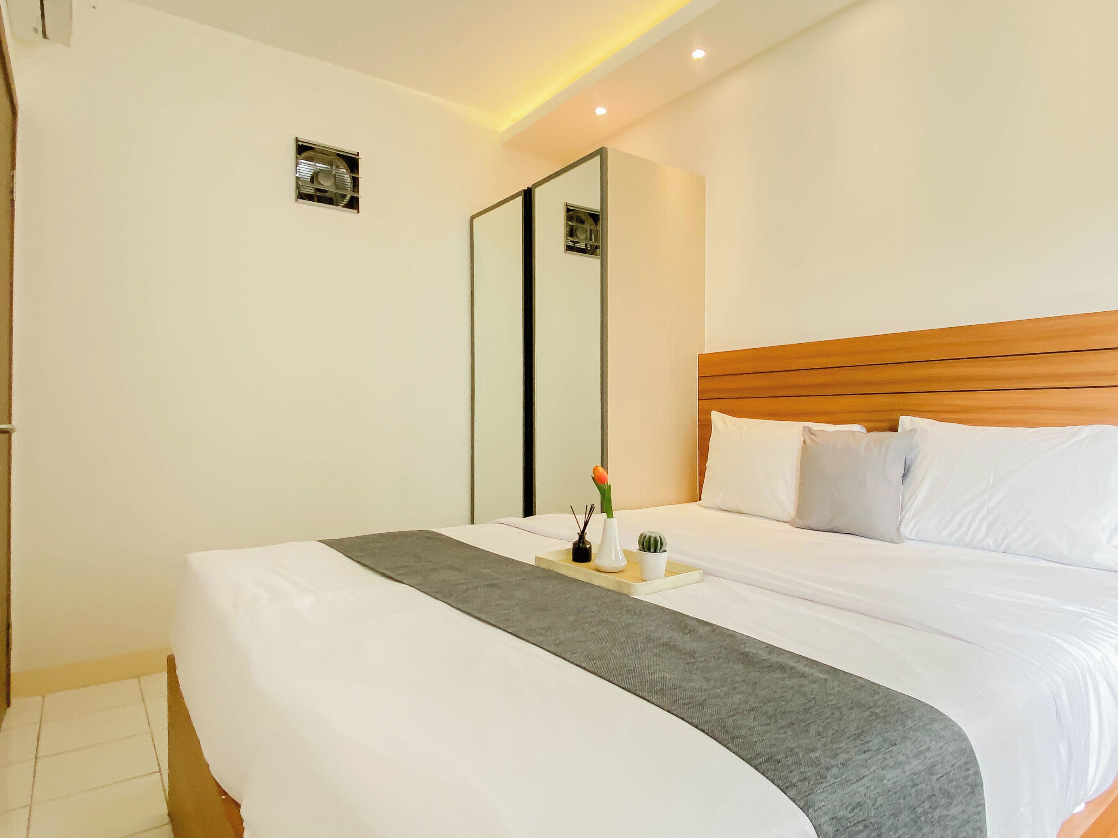A hotel room with a large bed, a wooden headboard, a mirror, and a bedside table with a vase and a small plant.