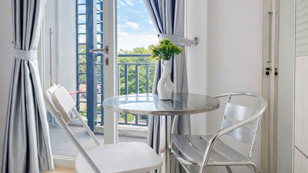 A balcony with a white chair, a small round table with a vase of green plants, and a view of trees and sky. - Classic White Studio At Apartment Woodland Park Residence (Jakarta)