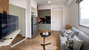 Un soggiorno moderno con una TV a schermo piatto, un divano grigio con cuscini decorati, un piccolo tavolo nero e un angolo cottura con frigorifero e fornello.
