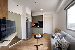 Ein modernes Wohnzimmer mit Flachbildfernseher, einer grauen Couch mit gemusterten Kissen, einem kleinen schwarzen Tisch und einer Küchenzeile mit Kühlschrank und Herd.