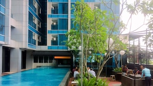 Una piscina con un edificio moderno al fondo, rodeada de vegetación y zonas de estar al aire libre.