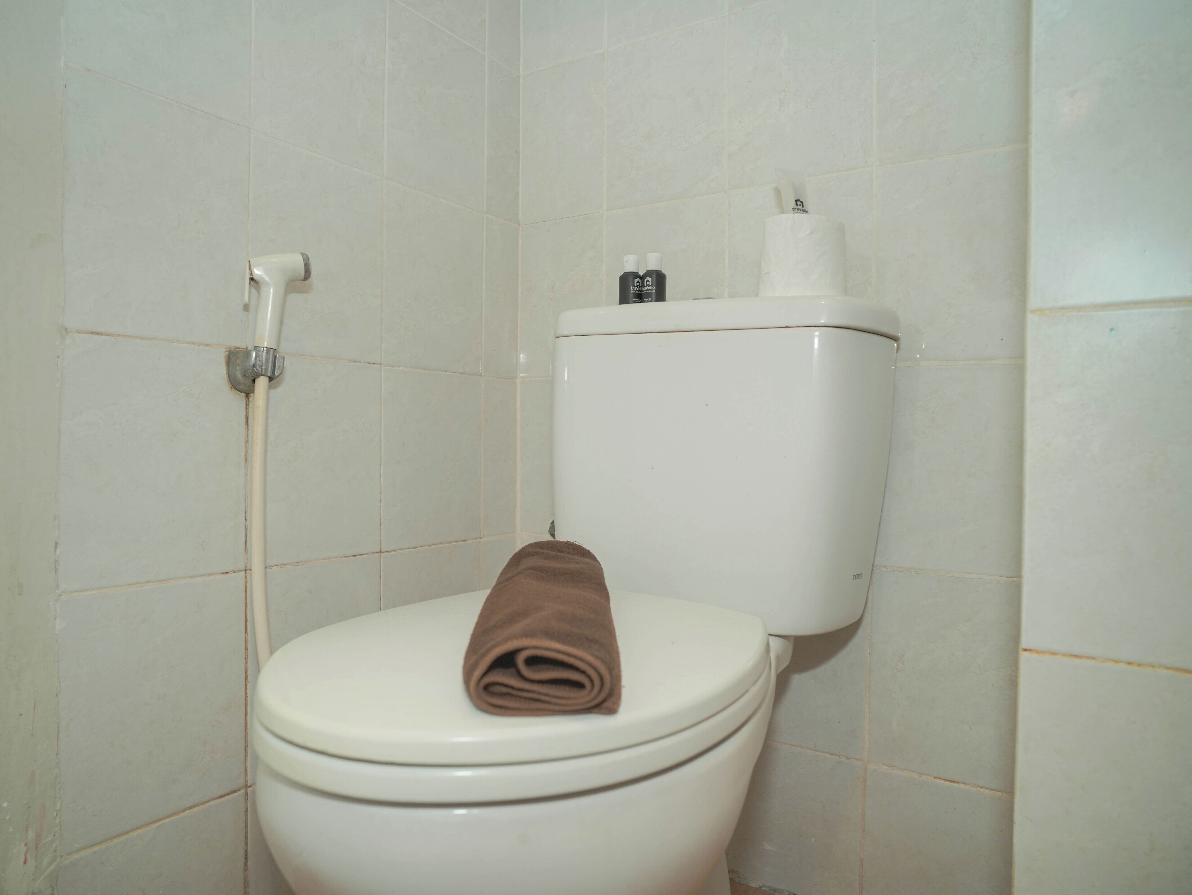 A toilet with a brown towel on the seat, a toilet paper holder, and a bidet spray on the wall.