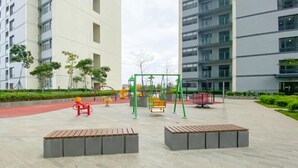 Ein Spielplatz im Freien mit Schaukeln, Rutschen und Fitnessgeräten, umgeben von befestigtem Untergrund und Grünflächen, mit modernen Gebäuden im Hintergrund.