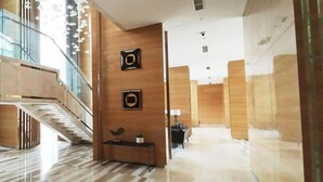 A modern hotel lobby with a wooden reception desk, a staircase, and a glass railing. - 6 Pax Tranquil 3Br With Extra Room At Grand Sungkono Lagoon Apartment (Surabaya)