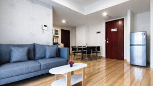 A modern living room with a sofa, a coffee table, a dining table with chairs, a refrigerator, and a door. - 6 Pax Tranquil 3Br With Extra Room At Grand Sungkono Lagoon Apartment (Surabaya)