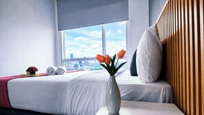 A hotel room with a bed, a window with a city view, a vase with flowers, and a bedside table. - 6 Pax Tranquil 3Br With Extra Room At Grand Sungkono Lagoon Apartment (Surabaya)