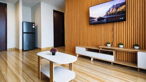A modern living room with a flat-screen TV mounted on a wooden wall, a white coffee table with a flower, a built-in TV stand with plants, and a kitchen area with a refrigerator. - 6 Pax Tranquil 3Br With Extra Room At Grand Sungkono Lagoon Apartment (Surabaya)