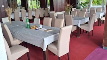 A dining area with multiple tables and chairs, a red carpet, and large windows with a view of greenery.