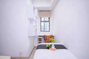 Quarto com cama, escrivaninha, cadeira e janela com vista para edifícios.