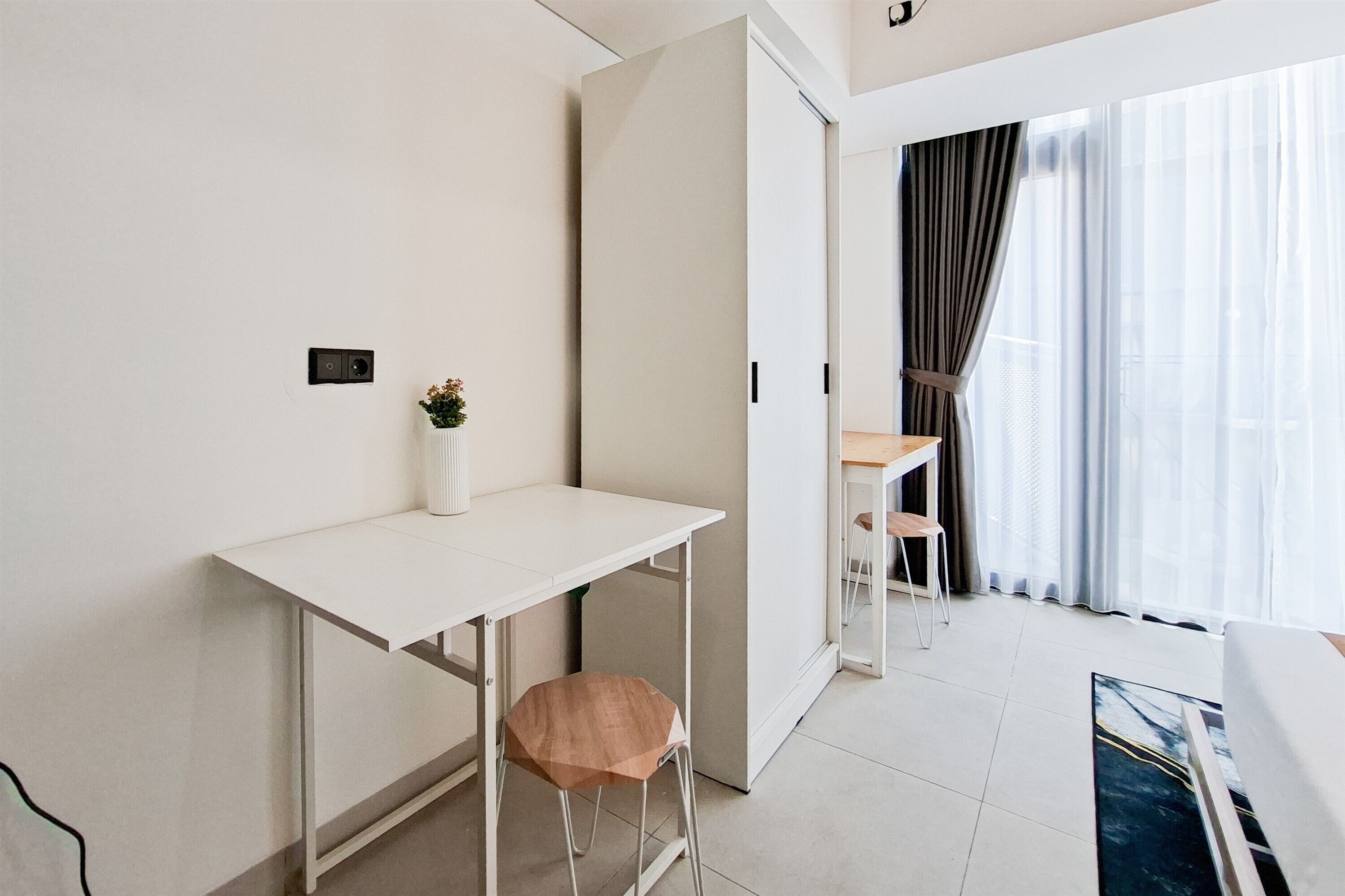 Ein modernes Zimmer mit weißem Schreibtisch, einer kleinen Hocker, einem Blumentopf, einer Schiebetür und Ausblick durch transparente Vorhänge.