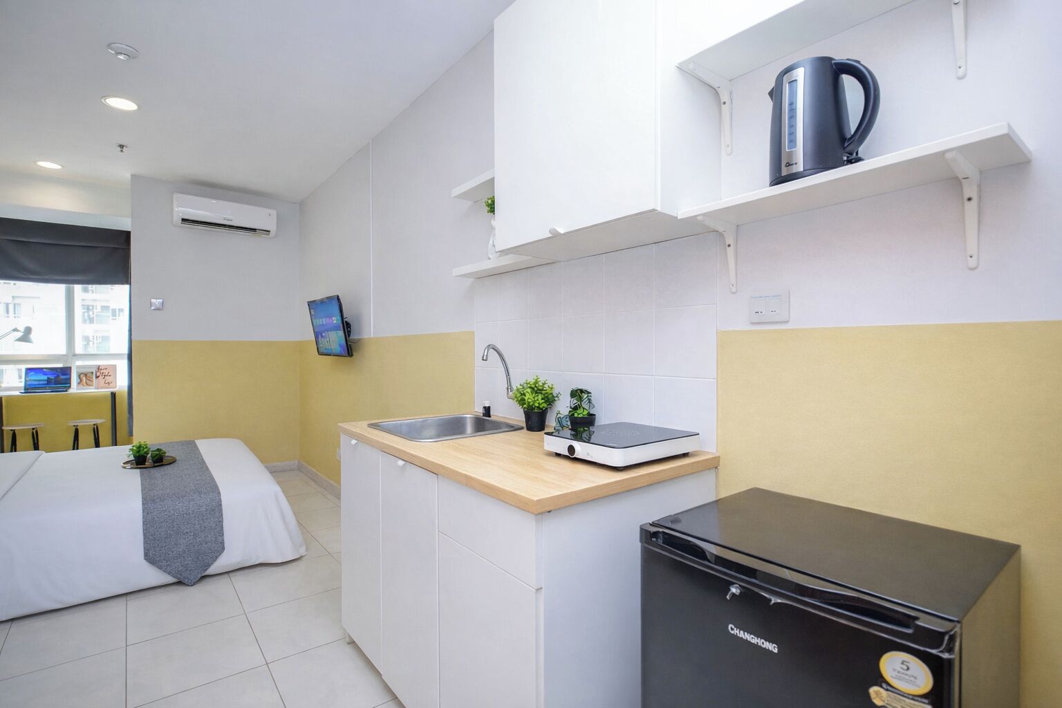 A compact hotel room with a bed, a small kitchenette, a black refrigerator, and a mounted TV.