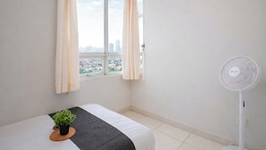 Une chambre d’hôtel minimaliste, comprenant un lit, une petite plante en pot, un ventilateur sur pied et une fenêtre avec des rideaux.