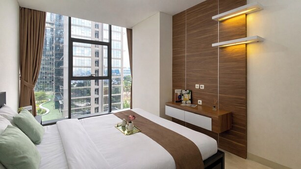Habitación de hotel con una cama grande, una mesita de noche, una lámpara de pared y una ventana con vistas a edificios.