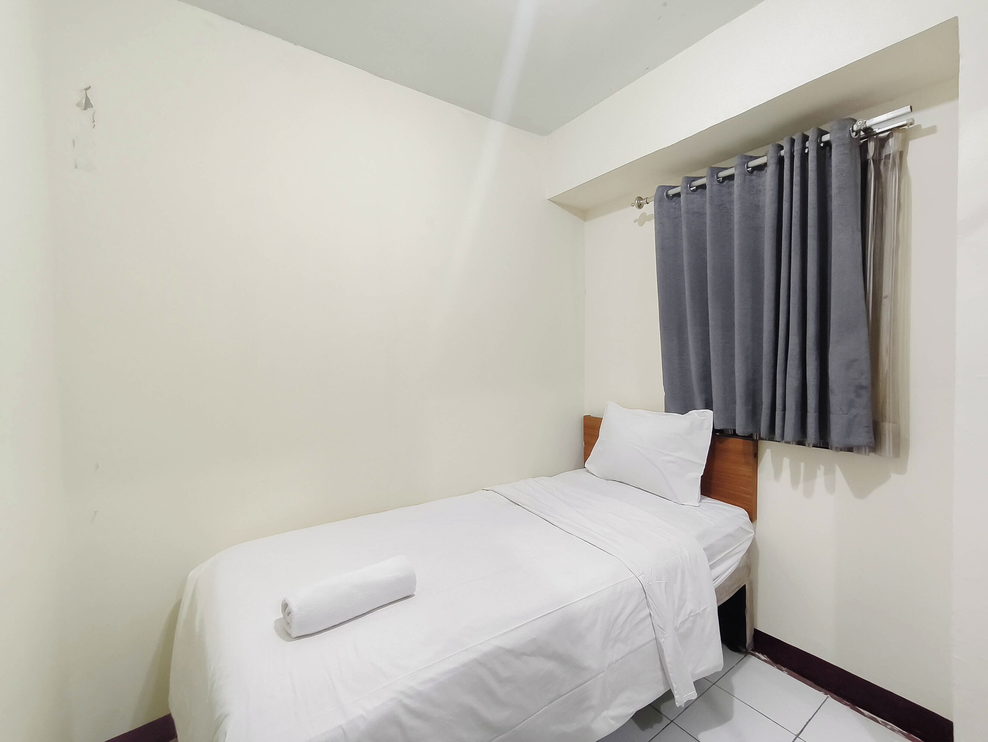 Una cama individual con colchón blanco y una toalla blanca enrollada, cabecera de madera, cortina gris y pared blanca.