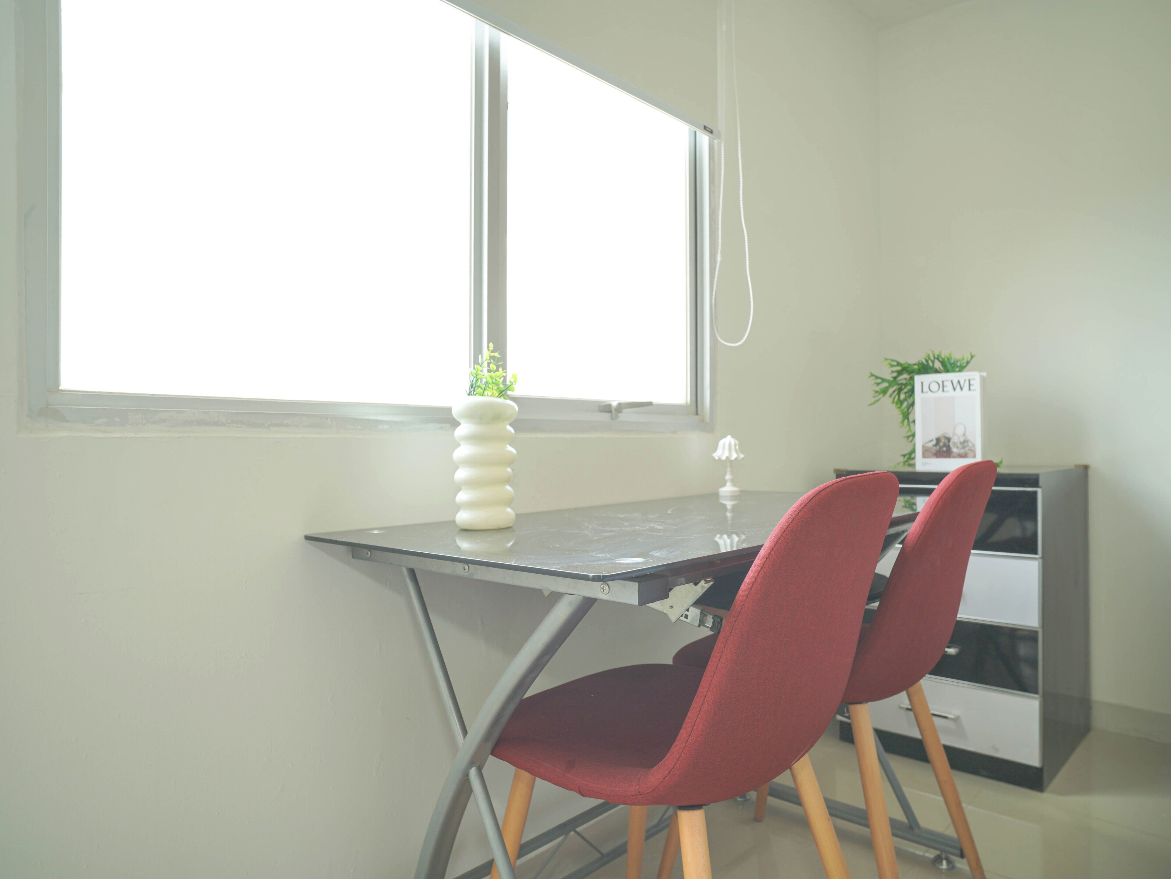 Een kleine kantoorruimte met een bureau, twee rode stoelen, een witte vaas met een plant, een boek getiteld 'LOEWE' en een raam met uitzicht.