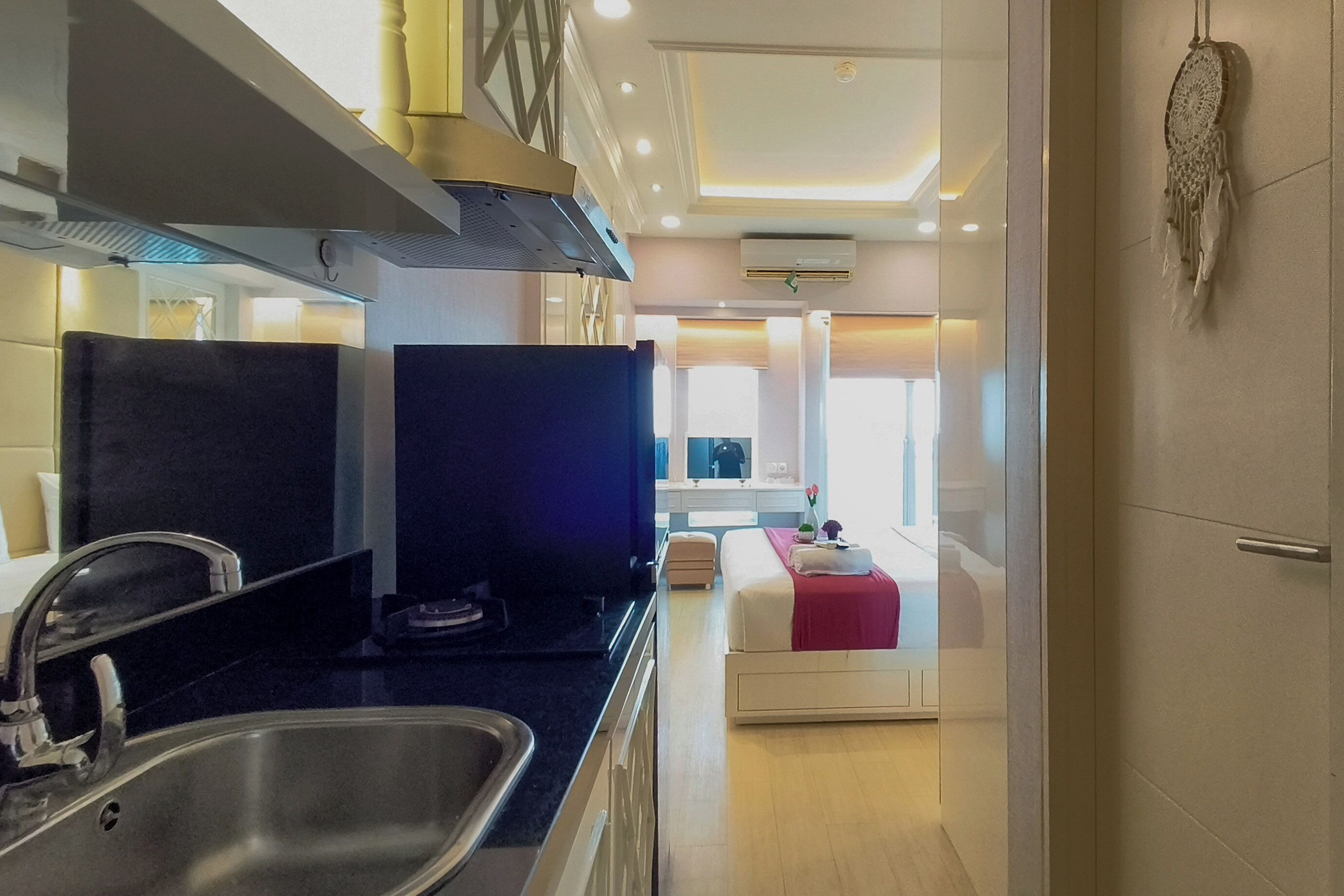 A modern hotel room with a kitchen area, a bed with a red blanket, and a dreamcatcher on the wall.