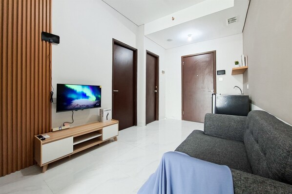 A modern living room with a flat-screen TV, a grey sofa, a wooden entertainment unit, and a kitchen area with a sink and cabinets. - Convenient And Roomy 2Br At Apartment Transpark Bintaro (South Tangerang)