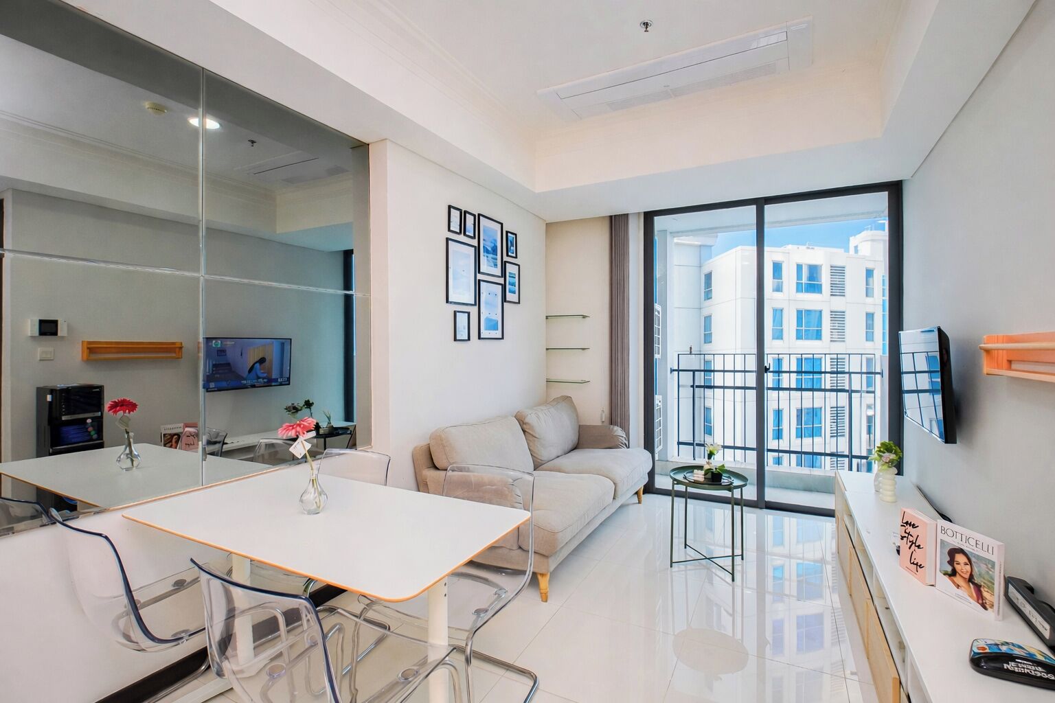 A modern living room with a sofa, a glass coffee table, a white dining table, and a large window with a view of buildings.