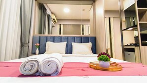 Quarto de hotel com cama, mesa de cabeceira com bandeja, planta em vaso e janela com cortinas.