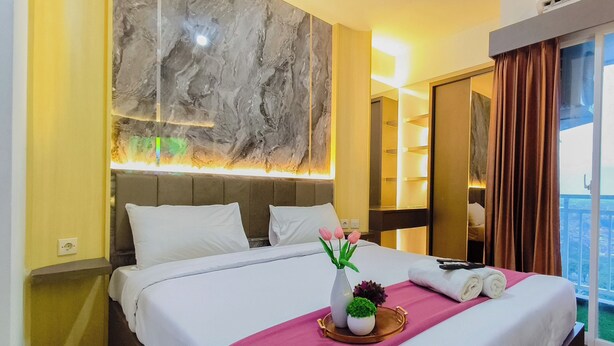 A hotel room with a bed, a wall-mounted light, a window with curtains, and a view of the outdoors. - Clean And Neat Deluxe Studio At Suncity Residence Apartment (Sidoarjo)
