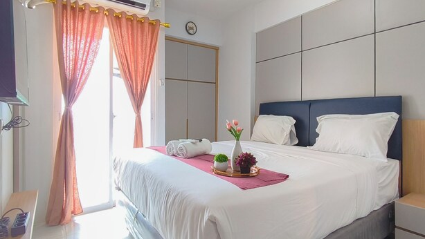 A hotel room with a bed, a bedside table, a nightstand, a window with curtains, an air conditioning unit, and a wall clock. - 2 Pax Practical Studio At Suncity Residence (Sidoarjo)