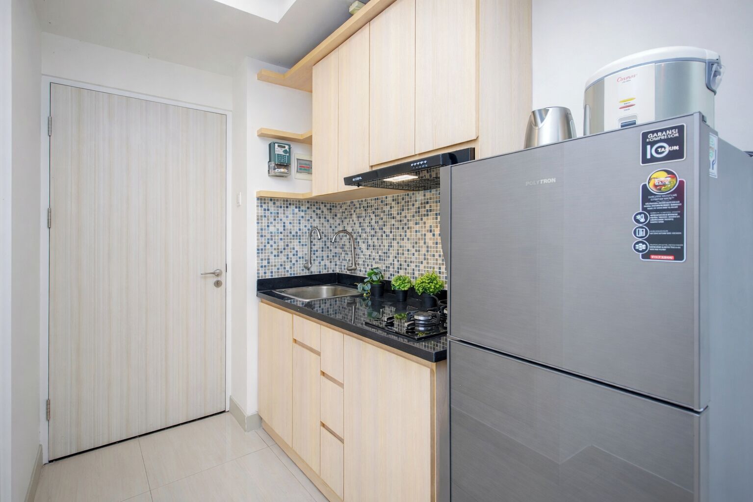 A compact kitchen with a stainless steel refrigerator, a tiled backsplash, wooden cabinets, and a small countertop with potted plants.