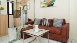 A living room with a brown sofa, patterned cushions, a glass coffee table, and a painting of leaves on the wall. - Practical 2Br For 3 Pax At Kebagusan City Apartment (Jakarta)