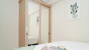 A bedroom with a bed, a bedside table with a tray, a green blanket, a mirror, and a wall-mounted plant. - Practical 2Br For 3 Pax At Kebagusan City Apartment (Jakarta)