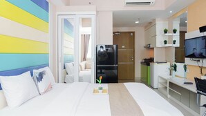A modern hotel room with a bed, a TV, a refrigerator, a desk, and a kitchenette. - Enjoy Restful Living In Studio Room At Apartment Oasis Cikarang (Cikarang)