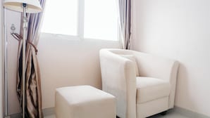 A room with a white armchair, a matching ottoman, a floor lamp, and a window with curtains. - Enjoy Restful Living In Studio Room At Apartment Oasis Cikarang (Cikarang)