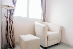 Um quarto com uma poltrona branca, um banquinho a combinar, um abajur e uma janela com cortinas.