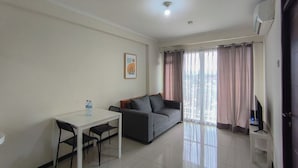 A living room with a sofa, a dining table with chairs, a TV on a stand, and a window with curtains. - Neat And Restful 2Br At Gateway Pasteur Apartment (Cimahi)