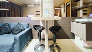 A modern living area with a sofa, two bar stools, a glass-top coffee table, a kitchen area with a sink, and a computer on a counter. - 4 Pax Warm Brown 2Br At Gateway Pasteur Apartment (Cimahi)