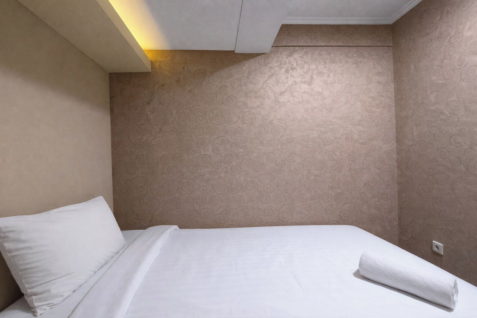 A hotel room with a bed, a pillow, a neatly folded towel, and a wall with a subtle pattern.