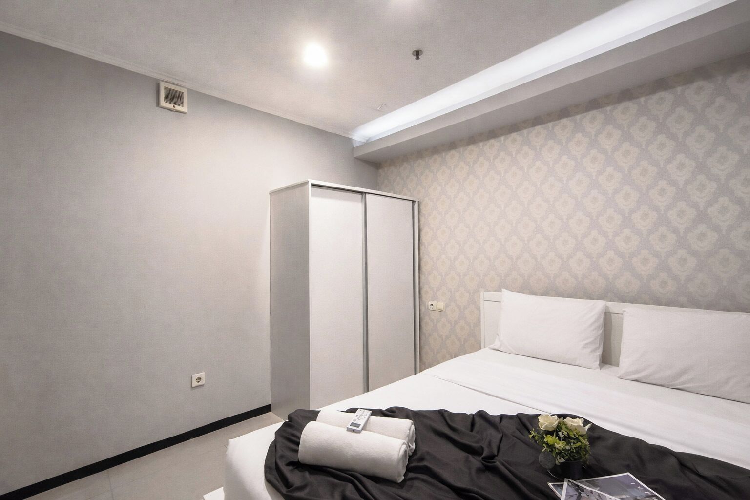 A hotel room with a bed, a wardrobe, a bedside table, and a wall with a patterned wallpaper.