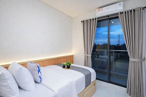A bedroom with a bed, pillows, a bedside table with plants, a window with curtains, and an air conditioning unit. - 2 Pax Practical Studio At Transpark Cibubur Apartment (Bekasi)