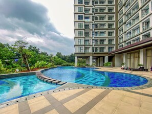 A swimming pool with a tiled deck, surrounded by greenery and a multi-story building with balconies. - 2 Pax Practical Studio At Transpark Cibubur Apartment (Bekasi)