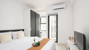 A hotel room with a bed, a small table with a tray, an air conditioning unit, a television, and a window with a view of buildings. - Bright And Comfy Studio Apartment At Transpark Cibubur (Depok)