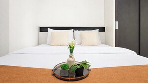 A hotel room with a bed, white bedding, two pillows, a bedside table with a tray holding a plant and a vase, and a dark wooden door. - Bright And Comfy Studio Apartment At Transpark Cibubur (Depok)