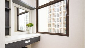 A bathroom with a white vanity, a mirror, a window with a view of a building, and a vase with flowers. - Bright And Comfy Studio Apartment At Transpark Cibubur (Depok)