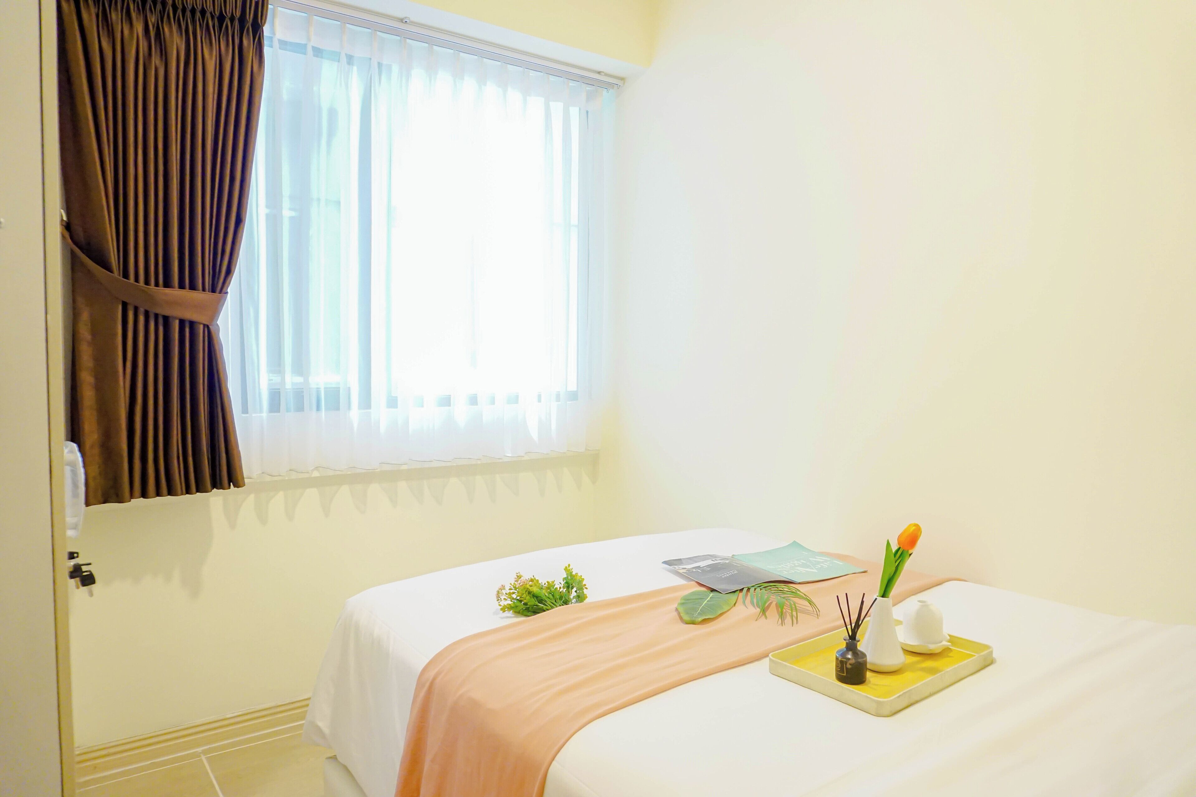 A hotel room with a bed, a window with curtains, a bedside table with a plant, a tray with a candle, and a small vase.