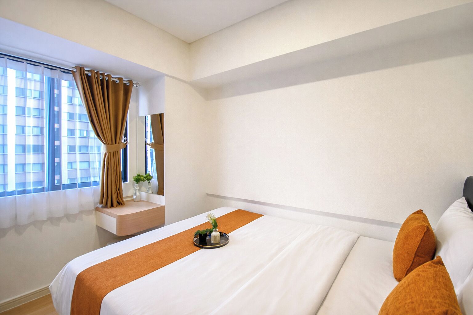 A hotel room with a large bed, a window with curtains, a small table with a vase, and a view of buildings outside.