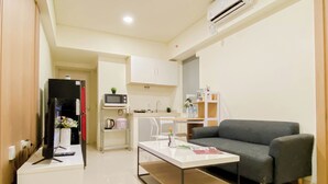 A modern kitchen with white cabinets, a microwave, and a sink. There's a grey sofa and a white coffee table with a magazine. - Good And Minimalist 2Br At Apartment Meikarta District 1 (Cikarang)