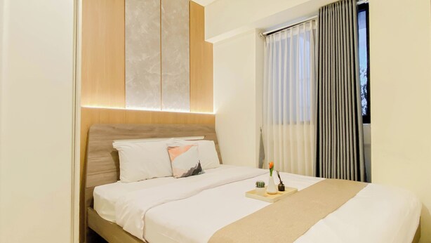 A hotel room with a bed, a bedside table, a window with curtains, and a wall-mounted air conditioner. - Good And Minimalist 2Br At Apartment Meikarta District 1 (Cikarang)