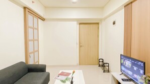 A modern living room with a sofa, a coffee table with a book and a flower, a TV on a stand, and a wooden door. - Good And Minimalist 2Br At Apartment Meikarta District 1 (Cikarang)