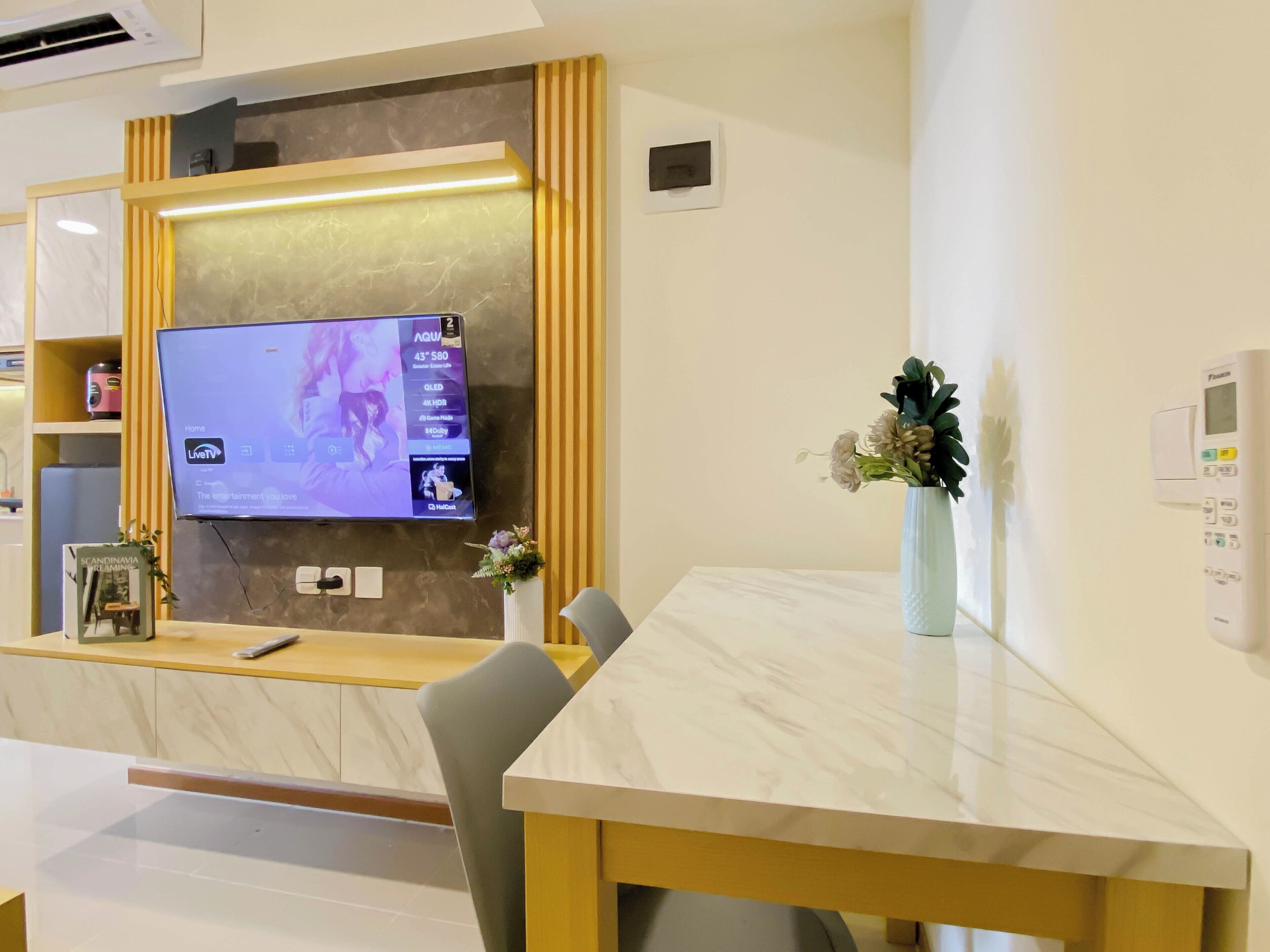 Zona de recepción de hotel con televisión de pantalla plana en la pared, un mostrador de recepción, una silla, un jarrón con flores y un termostato en la pared.