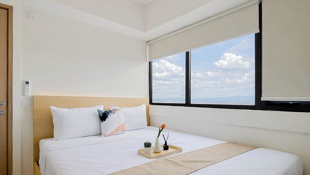 A hotel room with a bed, a window with a city view, a bedside table with a vase and a plant, and a door with a light switch.