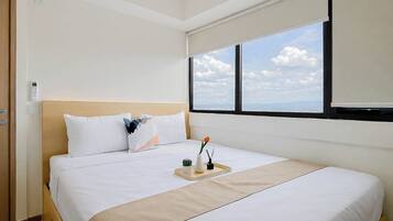 Habitación de hotel con cama, ventana con vista a la ciudad, mesita de noche con un florero y una planta, y una puerta con interruptor de luz.